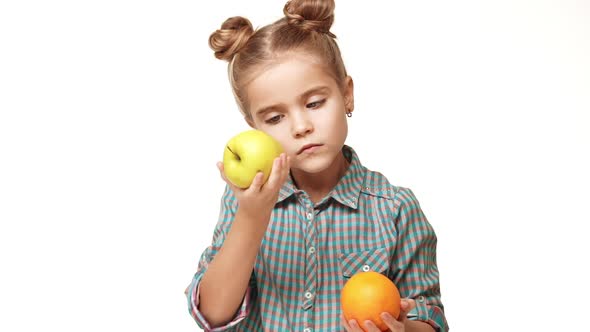 Young Smiling Small Beautiful Caucasian Girl Smelling Orange and Apple in Turns Than Choosing Apple