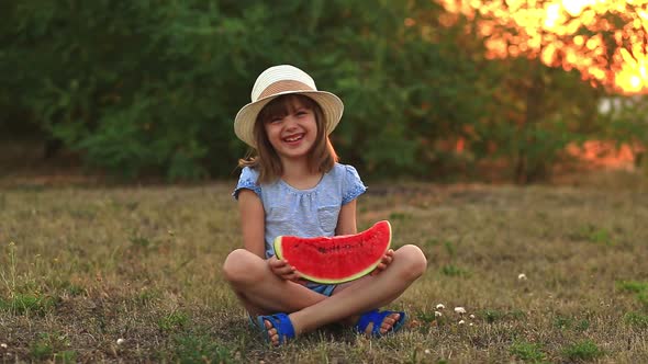 Funny Little Girl Smile and Eat Watermelon Slice Sitting on Grass with Crossed Legs in Summer Park