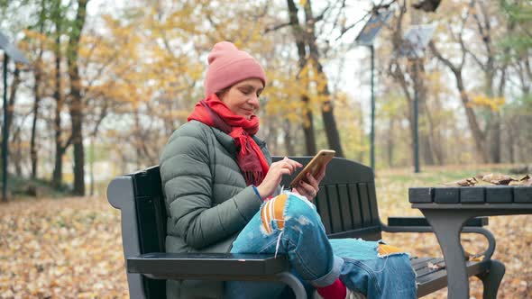 A Young Woman Using Her Smartphone While Sitting on the Bench in the Autumn Park