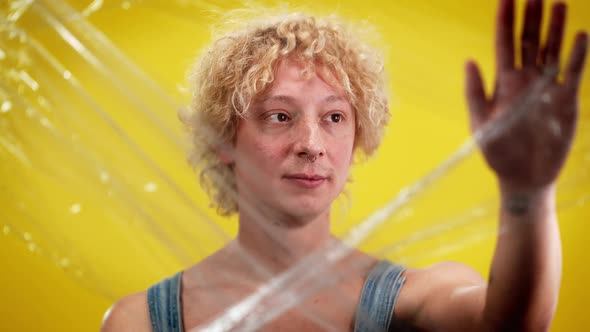 Portrait of Androgynous Caucasian Young Man Touching Transparent Film Standing at Yellow Background