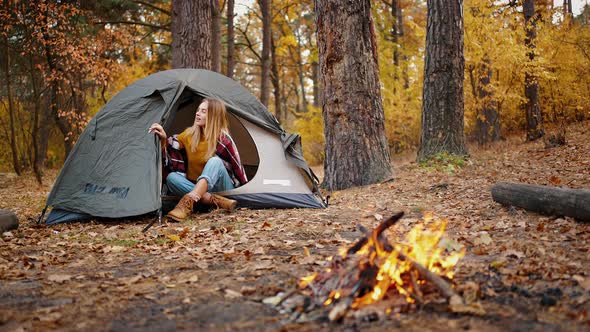 Young Woman Sitting in Tent Looking Out of It and Smiling Rejoicing in the Beauty of Autumn Forest