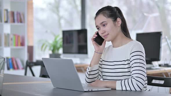 Young Asian Woman with Laptop Talking on Smartphone in Office