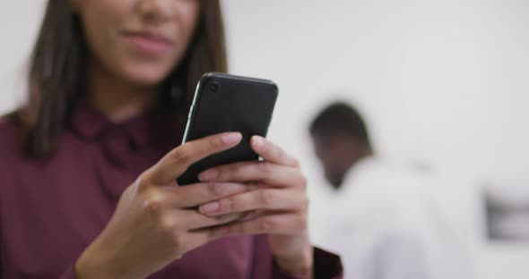 Smiling biracial businesswoman using smartphone with colleagues in background in modern office