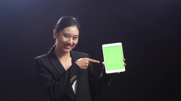 Asian Speaker Woman In Business Suit Holding And Pointing Green Screen Tablet In The Black Studio