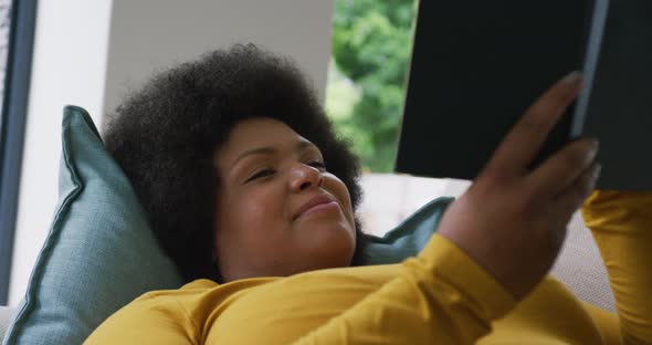 Video of happy plus size african american woman lying on sofa and reading book