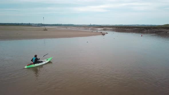 Man kayaking on meandering river past a colony of seals, with flocks of birds flying in the backgrou