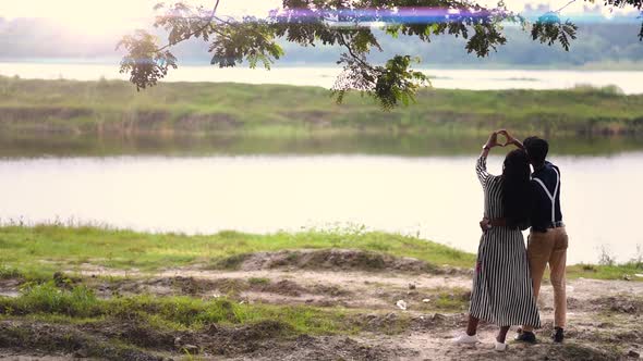 Romantic couple in love making heart shapes with hands, near a lake in forest at sunset or sunrise