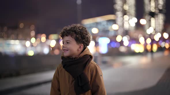 a Curlyhaired Smiling Boy Stands on an Evening Street Against the Background of Blurred City Lights