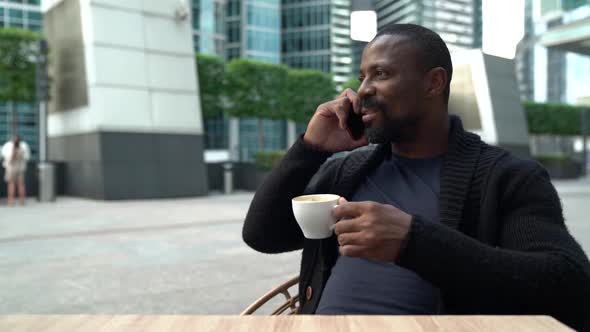 Black Man Is Relaxing in Cafe on Street in Break, Drinking Tea or Coffee and Calling By Phone