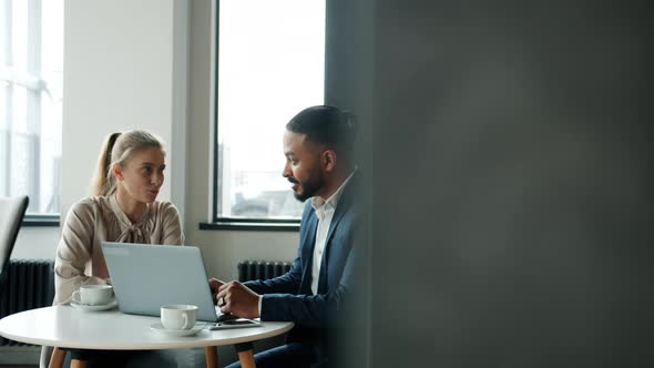 Arab Man and Caucasian Woman Talking and Working with Laptop Sitting at Table with Coffee