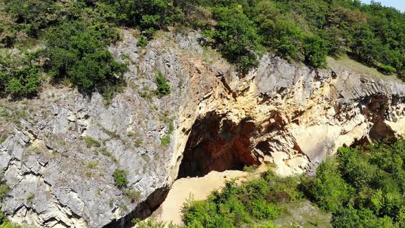 Open cave in rocky hill, all covered with green bushes on top, aerial view