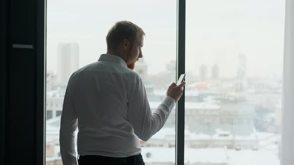 online businessman in smartphone stands by the window with city view