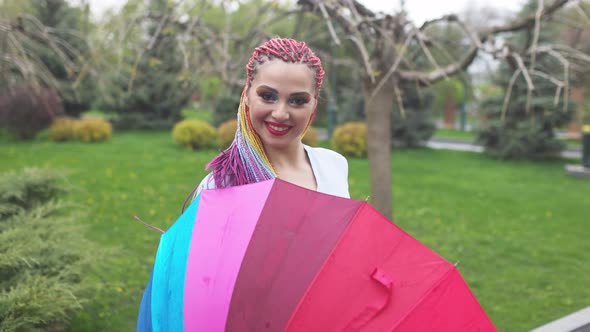 A Cute Girl with Multicolored Braids and Bright Makeup in a Bluish Shirt Posing with a Rainbow