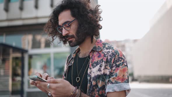 Concentrated curly-haired bearded man in eyeglasses looking at phone and around