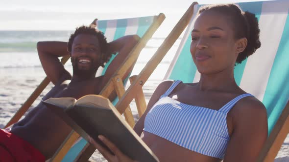 African american couple sunbathing and reading book while sitting on deck chairs at the beach