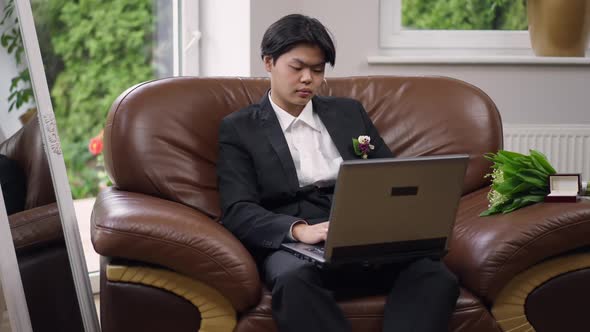 Concentrated Asian Young Man in Wedding Suit Surfing Internet Typing on Laptop Keyboard with Serious