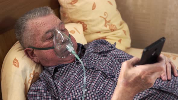 Old Man Wearing Breathing Mask Is Laying in Bed Holding Smartphone