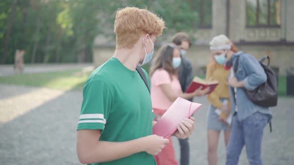 Young Redhead Man in Face Mask Looking Back at Groupmates and Turning To Camera. University Students