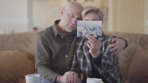 Adult Couple Sitting on a Couch Examines Their Old Photos and Shares Their Memories