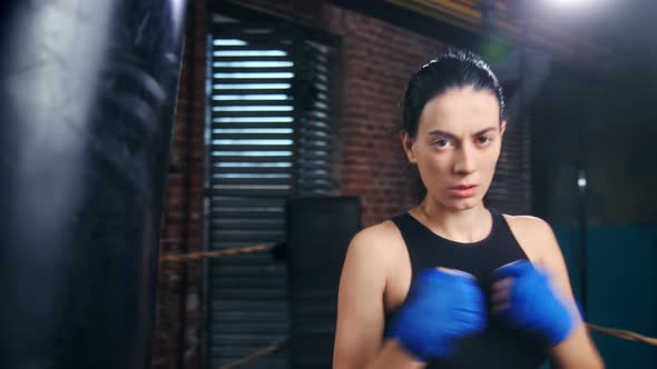 Aggressive Female Kickboxer with Drops of Sweat on Face in Blue Wrapped Hands Posing at Dark Gym