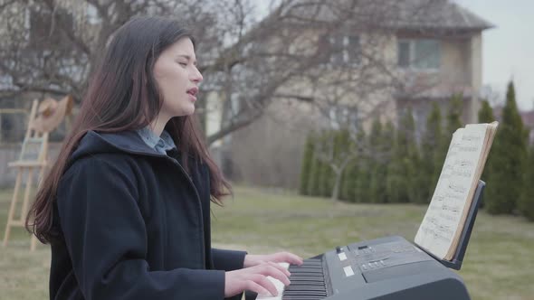 A Successful Beautiful Brunette Girl Plays on a Synthesizer and Sings While Sitting in the Backyard