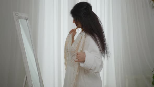 Side View of Slim Beautiful Young Woman in Bathrobe Standing in Front of Mirror Trying on Wedding