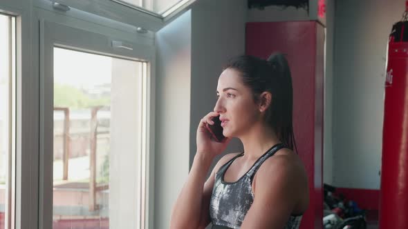 Young Woman in Sport Outfit is Standing Near the Window Talking on the Phone and Drinks Multivitamin