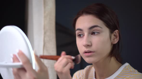 A Young Woman Looks Into a Small Mirror and Applies Makeup with a Brush. Skin Care.