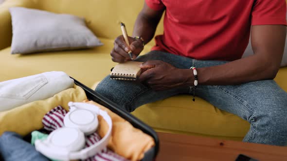 Young Adult Man Writing Travel Checklist While Packing Clothes to the Suitcase