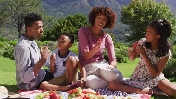 Happy african american parents, daughter and son sitting outdoors on blanket, eating fruits