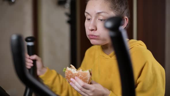 girl idly trains on a sports machine and chews a burger
