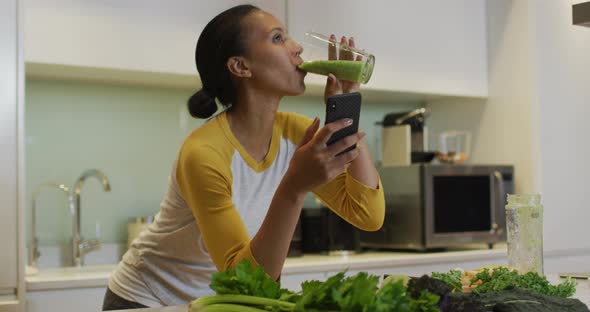 Mixed race woman drinking healthy drink and using smartphone in kitchen