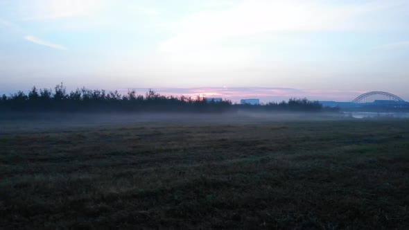 flying over the grass where the fog is hanging on the ground with a bridge, buildings and sunrise on