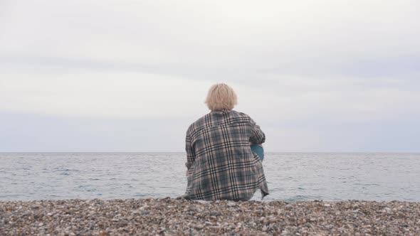 A Young Man in a Plaid Shirt Sits on the Shore and Throws Stones Into the Sea