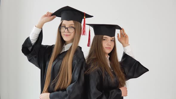 Two Young Happy Graduate Women Stand Shoulder to Shoulder Correct Their Hats with Their Hands