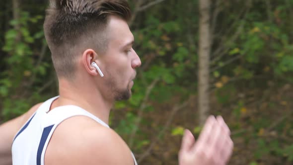 Athletic Sporty Guy with Wireless Headphones Sprinting Fast Along Trail Near Forest. Young Strong