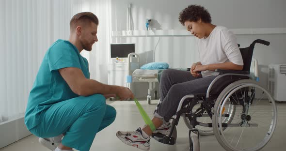 Male Nurse Supporting Disabled African Woman Leg in Wheelchair