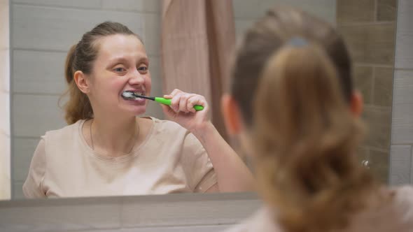 Woman in Beige Tshirt Actively Brushes Her Teeth with Green Toothbrush in the Bathroom at Home