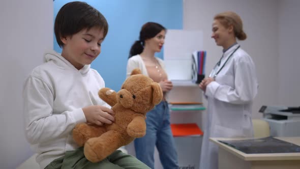 Side View Positive Caucasian Son Playing with Teddy Bear As Blurred Mother Talking to Pediatrician