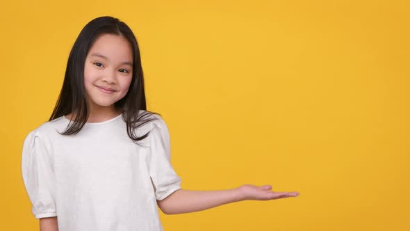 Adorable Little Asian Girl Outstretching One Hand Aside Demonstrating Something Over Orange Studio