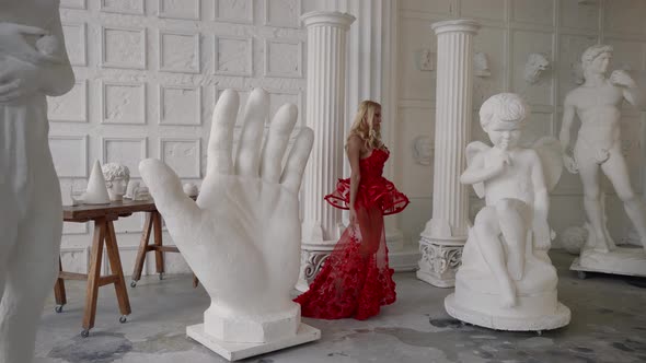 Sexy Blonde Lady in Red Dress is Walking in Workshop of Sculptor Between Sculptures