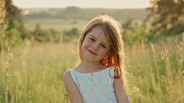 Portrait of a Cute Girl with Flowing Hair in a White Summer Dress Standing on a Green Lawn Holding