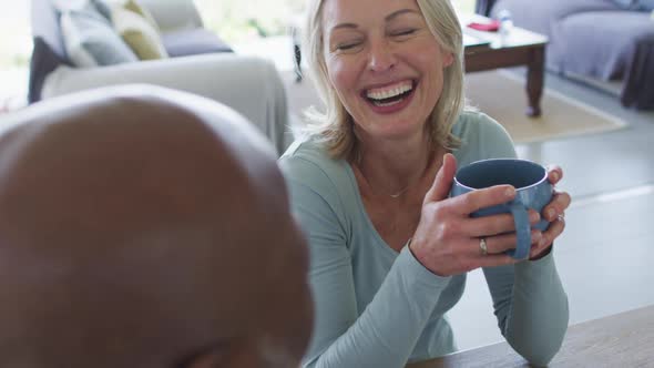 Caucasian senior woman holding a coffee cup smiling while talking to her husband at home