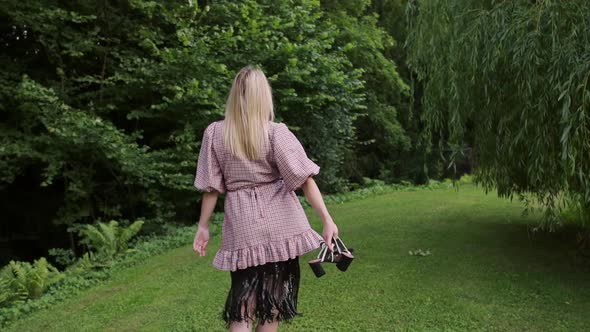 Fashion woman holding shoes in hand walking on grass through green public garden