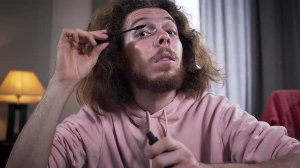 Headshot of Caucasian Intersex Person Applying Mascara and Smiling. Man Doing Makeup on One Side of