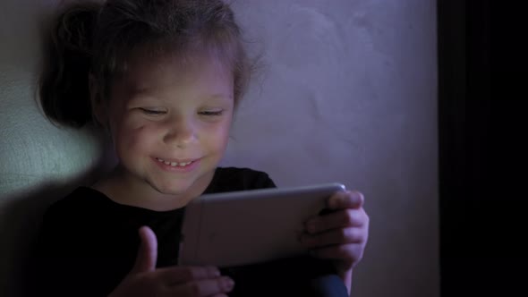 Little Girl Sitting on the Floor and Playing Tablet or Smartphone in Social Internet in Dark Blue