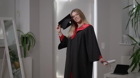 Smiling Beautiful Grad Woman Posing with Rolled Certificate and Graduation Cap Indoors at Home