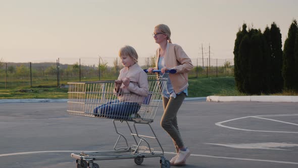 Woman Having Fun in the Parking Lot at the Supermarket, Rolls the Child in the Shopping Trolley