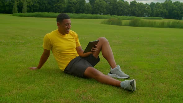 Cheerful Relaxed Sporty African Man Watching Fitness Video on Tablet Pc in Park