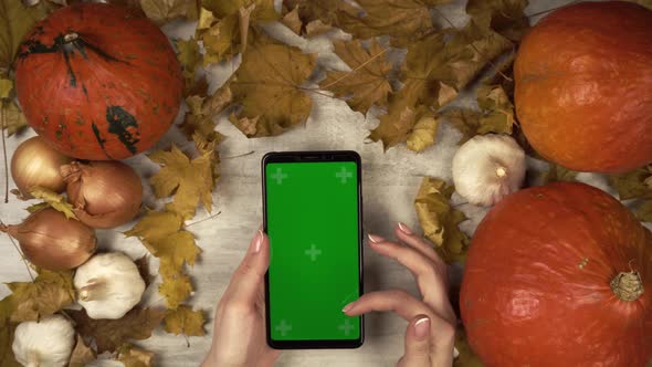 Woman Clicks on a Green Screen Smartphone in Vertical Orientation in the Middle of Fall Decoration
